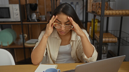 Woman stressed in apartment living room with laptop and notepad, feeling overwhelmed while...