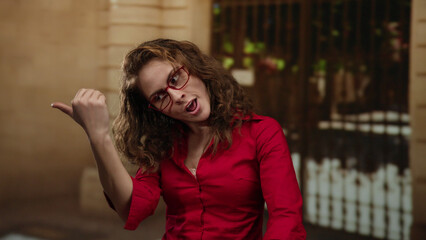 Woman gestures energetically while wearing red and glasses on a city street outdoors, showcasing lively expression in an urban european landscape.
