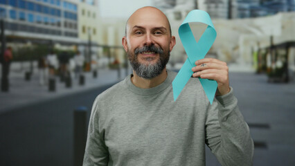 Caucasian man with bald head and beard holds blue cancer awareness ribbon on urban street...
