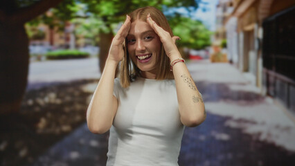 Fototapeta premium Young woman with tattoos playfully covers her face while standing on a sunny urban street, surrounded by vibrant city life and green trees, embodying a cheerful and carefree spirit.