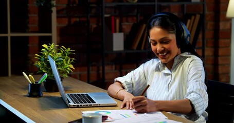 Indian businesswoman taking notes from video call on laptop using headphone at desk in modern cozy office, Asian female corporate executive jotting down important points attending virtual meeting - Powered by Adobe