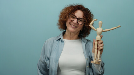 Woman holding wooden mannequin smiles against isolated blue background showcasing artistic expression and creativity in a minimalist setting.