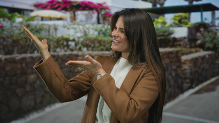 Young hispanic woman smiling and pointing with hands on a vibrant city street surrounded by colorful flowers and outdoor settings wearing a stylish coat in the afternoon light.