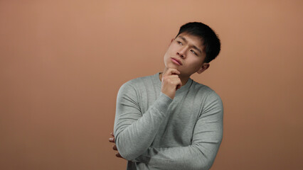 Young man in gray shirt over a brown background with a thoughtful expression, exemplifying contemplation and introspection in a studio setting.