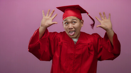 Young man in red graduation gown making playful faces against pink wall, celebrating graduation day.