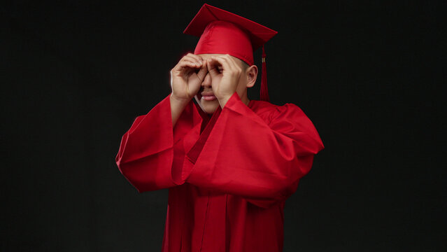 Young chinese man in red graduation cap and gown makes binocular gesture isolated on a black background, symbolizing curiosity and future vision.