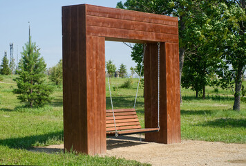 A wooden swing bench suspended by chains within a modern park installation surrounded by lush greenery. Photo