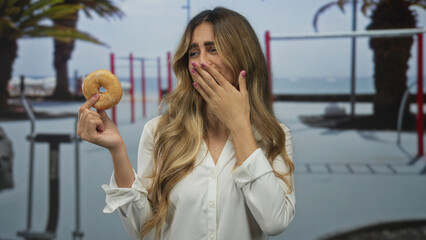 Young blonde woman holds donut with one hand and covers mouth for disgust inside gym; refusal...