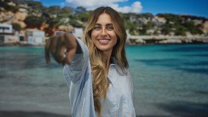 Young blonde woman with long hair pointing finger directly at camera on sunlit beach; invitation...