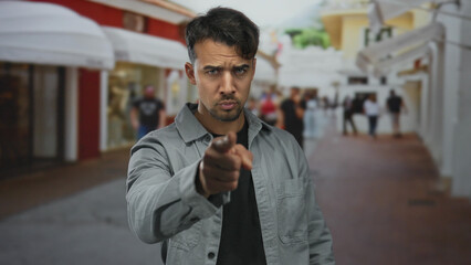 Obraz premium Young hispanic man gestures on a bustling city street, inviting the camera with a confident expression, surrounded by urban outdoor scenery and blurred passersby.