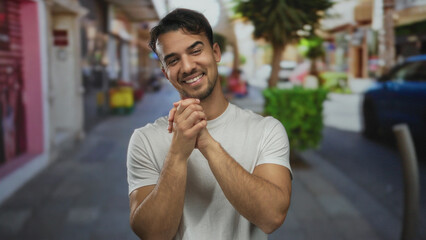 Young hispanic man in white shirt showing gratitude on a vibrant city street outdoors.
