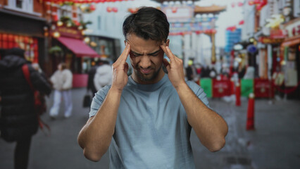 Young hispanic man feeling stressed in bustling city street with colorful decorations blurred in...