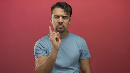 Young hispanic man in blue shirt pointing finger seriously against isolated red background showing...