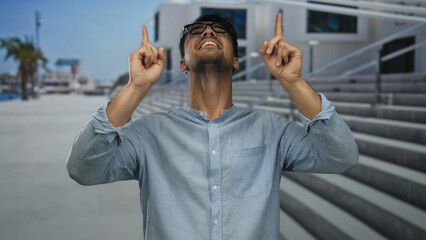 Young hispanic man outdoors pointing upwards with a joyful expression near modern architecture and...