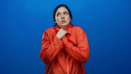 Woman in an orange prison jumpsuit, with brunette hair, looking cold against a blue background,...