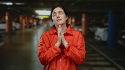 Woman in orange prison jumpsuit with handcuffs looks pleading in a dimly lit parking area with cars...