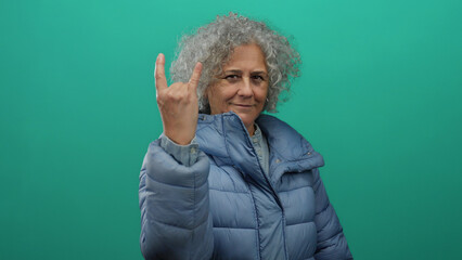Woman with curly grey hair in a blue jacket making a rock gesture against a green background,...