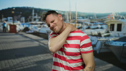 Fototapeta premium Young man in a striped shirt at a seaside port holding his neck in discomfort, with boats in the background and a clear sky, conveying a nautical theme and relaxed setting.
