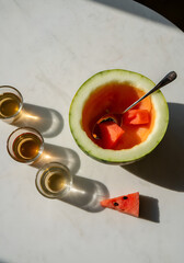 Slices of watermelon with visible seeds, small glass cups with water or tea, and a reflective spoon resting inside a hollowed melon half, arranged on a sunlit white marble tabletop during golden hour.