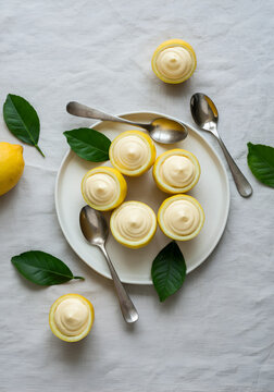 A natural gourmet food scene featuring lemon halves filled with creamy lemon mousse, arranged on a white ceramic plate with fresh green leaves and silver spoons