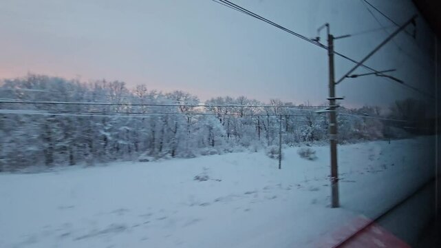 Train window shows a snowy landscape with trees and wires. The sky is dark blue and the sun is setting. view from moving train commuter transport passenger express.