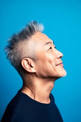 Fototapeta premium Creative pattern of style for aging men. A joyful and radiant male expressing confidence. Blue solid background. Trendy men's crest haircut - midlife asian man with middle hair.
