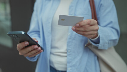 Woman holding smartphone and credit card while standing on an outdoor street, wearing a blue shirt, suggesting online shopping or mobile banking.