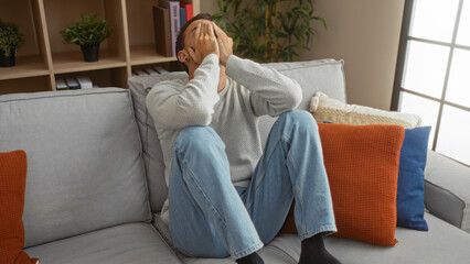 Young man sitting on a couch in a living room with his hands covering his face, surrounded by...