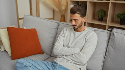 Young hispanic man sitting on a couch with arms crossed indoors in a modern apartment living room...