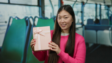 Woman on bus in pink sweater holding gift smiling indoors, showcasing a sense of celebration and...