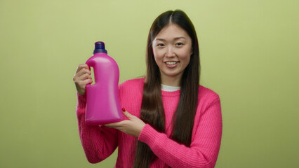 Woman holding detergent bottle wearing pink sweater smiling against yellow background in studio...