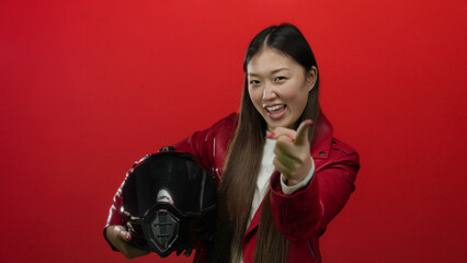 Woman pointing with a playful gesture holding helmet against vibrant red background, wearing...