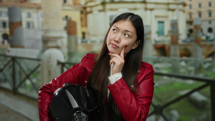 Woman in red jacket holds helmet while pondering in front of ancient roman ruins in city setting, combining modern and historical elements outdoors.