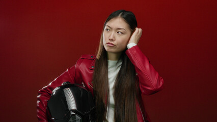 Woman wearing red jacket holding helmet in front of red background appearing thoughtful and focused
