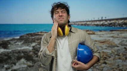 Hispanic man wearing headphones and hardhat stands on a beach holding his cheek in discomfort, with...