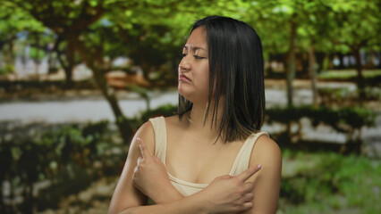 Young chinese woman outdoors in a park crossing her arms with a displeased expression while standing under green trees reflecting uncertainty.