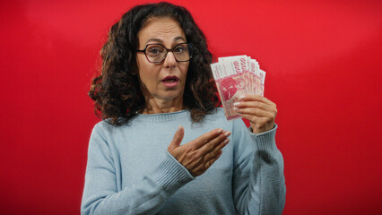 Woman holding icelandic banknotes on isolated red background, expressing curiosity with middle age...