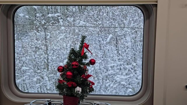 MOSCOW - DEC 25, 2023: Small Christmas tree with red ornaments sits in a window. The tree is surrounded by snow in train restaurant dining car railroad transit connection on ground