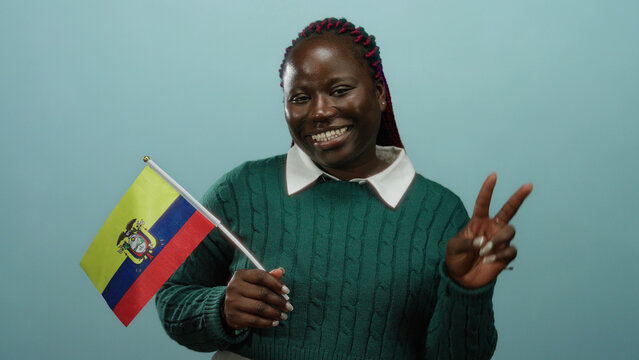 Woman with braids smiles holding colombia flag against blue wall, gesturing victory, conveying national pride.