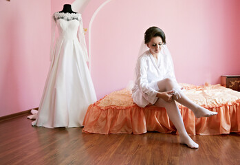 A bride is getting ready for her wedding and is sitting on a bed. A wedding dress is in the...