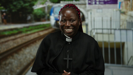 Woman laughing in priest attire at a train stop with blurred train in background, expressing joy...