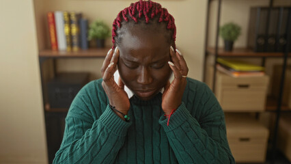 Woman with braids feeling stressed in an office setting with books and plants in the background, emphasizing her emotion and indoor work environment.