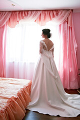 A woman in a white wedding dress stands in front of a pink curtain. She is looking out the window,...
