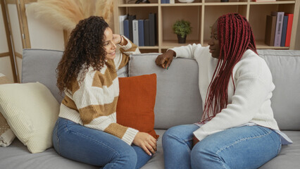 Two women sitting on a couch in a cozy living room enjoying a friendly conversation together, highlighting warmth and connection in a relaxed indoor setting.