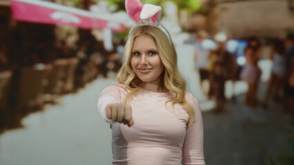 Fototapeta premium Young blonde woman wearing bunny ears playfully greets with a fist bump on a sunny city street, embodying joyful outdoors interaction with a humorous twist.
