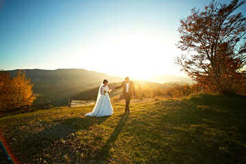 A bride and groom are walking in a field with a beautiful sunset in the background. Scene is...