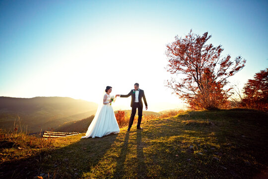 A bride and groom are walking together on a grassy hill, with the sun shining brightly on them. The bride is wearing a white dress and holding a bouquet, while the groom is wearing a suit