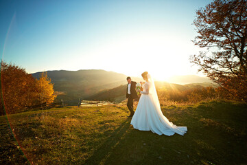 A bride and groom are walking together in a field with a beautiful sunset in the background. The...