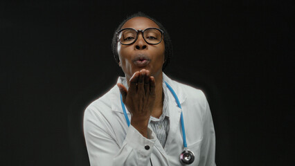 Woman doctor in white coat with stethoscope sending kiss on black background shines in professional...