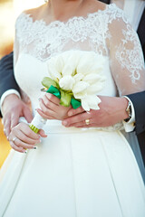 A bride and groom are holding hands and a bouquet of white flowers. The bride is wearing a white...